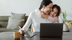 Mother and daughter hugging at table while mother is at computer..