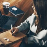 Woman write on notepad next to laptop with airpods and a cup of coffee.